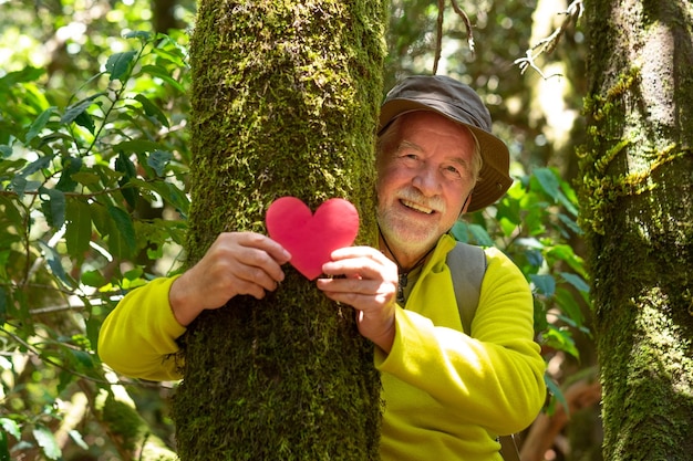 Smiling senior man partially hidden by a moss covered tree trunk in the woods holding a paper heart in his hands earth day concept