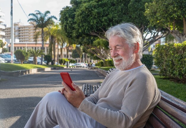 휴대 전화를 사용하여 공공 공원의 벤치에 앉아 웃고 있는 노인 - 기술과 사교를 즐기는 매력적인 흰 머리 노인