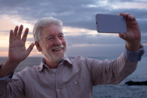 Smiling senior man standing in front of the sea in video chat by his mobile phone while the sun sets in the sea Peace relaxation and beauty in nature