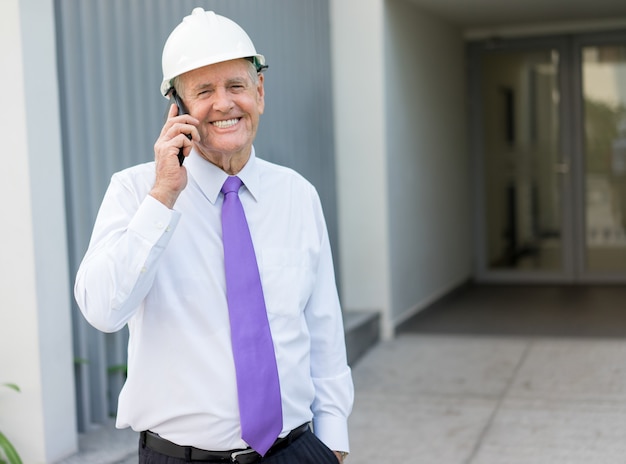 Smiling senior man in tie and helmet talking on phone at construction site. 