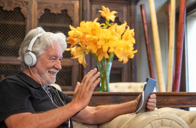 Smiling senior man using electronic device with headphones in video call. Sitting inside in armchair