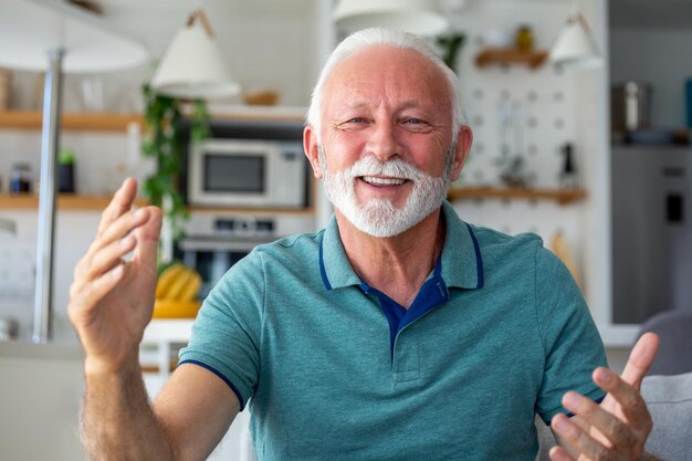 写真 笑顔の高齢男性がカメラに向かって手を振りラップトップでビデオコールをしている幸せな高齢の男性が自宅のソファに座って近代的な技術とワイヤレス接続を使用して話します