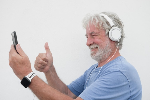 Smiling senior man wearing headphones in video call with phone and thumb up copy space attractive bearded joyful senior male enjoying tech and social