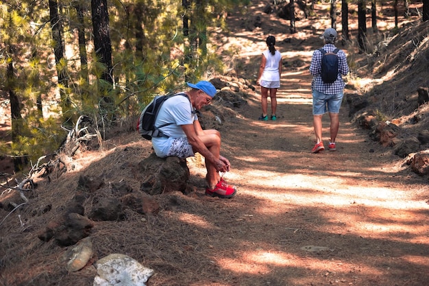 Smiling senior man with backpack on shoulders stops during mountain hike to tie his shoes