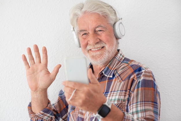 Smiling senior man with white hair using mobile phone in video chat wearing headphones. Gesturing a greetings with hand