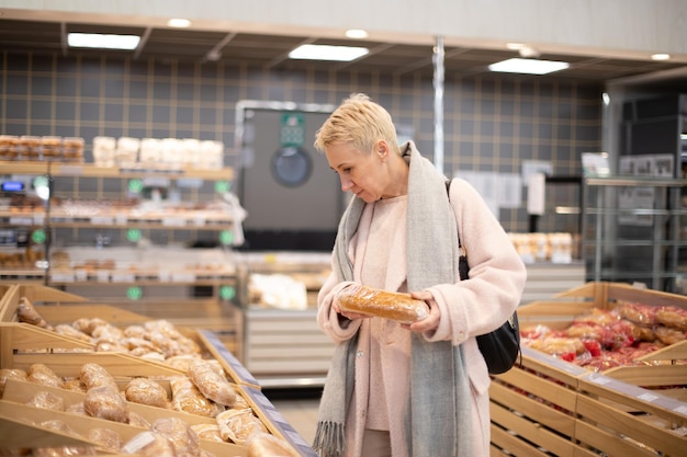 Улыбающаяся пожилая зрелая женщина выбирает хлеб и выпечку в продуктовом отделе супермаркета в пекарне