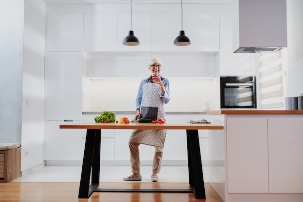 Smiling senior woman in apron standing in kitchen, drinking wine and preparing healthy dinner