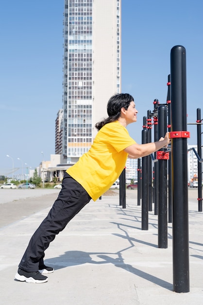 Улыбающаяся старшая женщина делает отжимания на открытом воздухе на барах спортивной площадки