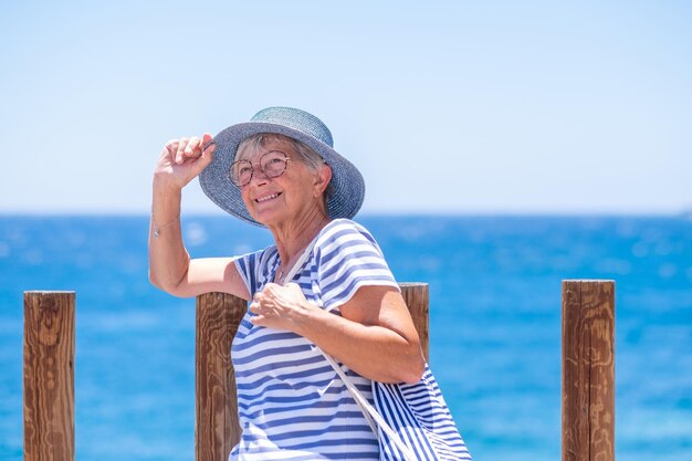 Smiling senior woman dressed in blue with blue hat sitting close to the sea in summer vacation enjoying sunny day