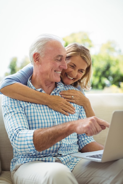 Donna senior sorridente che abbraccia un uomo in salone mentre per mezzo del computer portatile