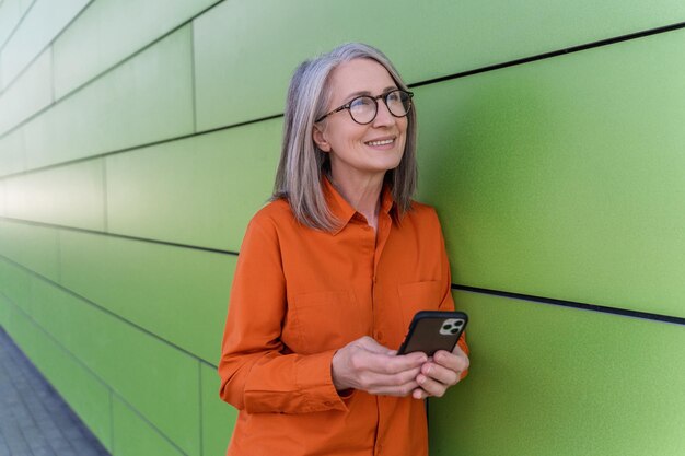 Smiling senior woman holding smartphone checking mobile app online shopping standing on the street