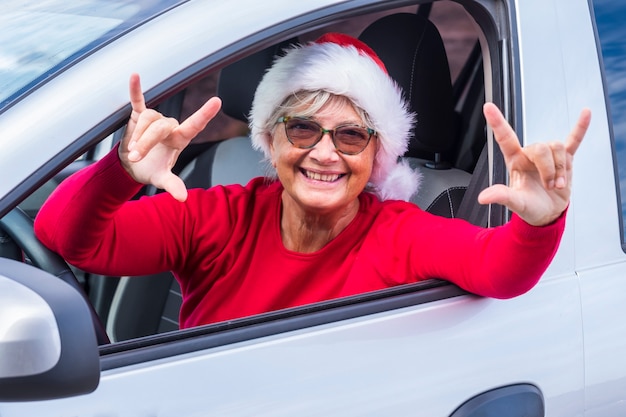 笑顔の年配の女性は、クリスマスの帽子をかぶって車の窓から身を乗り出し、次の休日を待っている彼女の手で前向きな兆候を身振りで示す笑顔-イベント