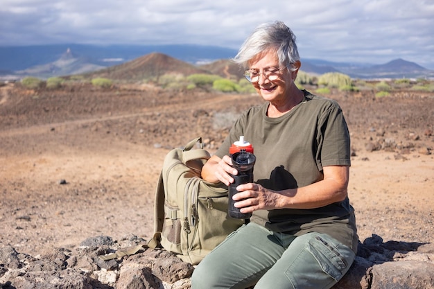 Smiling senior woman on outdoor hike sitting on a stone wall holding her water bottle Active mature woman sitting near her backpack enjoying freedom and sunny day