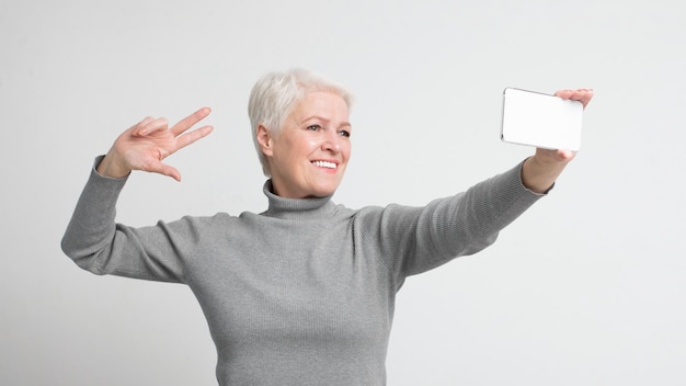 Photo smiling senior woman posing with smartphone and peace sign