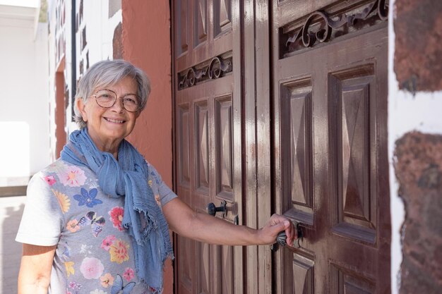 Smiling senior woman short hair wearing eyeglasses and scarf opening a large wooden door of the country house when come back home