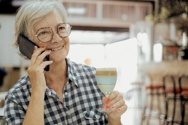 Smiling senior woman sitting at cafe table talking using mobile phone holding a drink glass