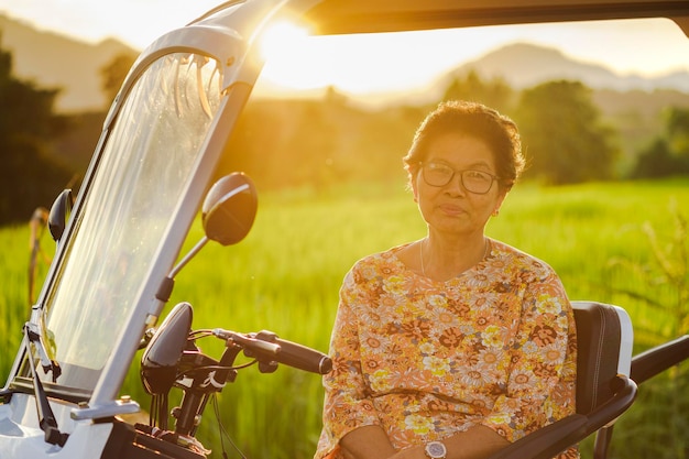 Smiling senior woman sitting on electric tricycle with rice fields and sunlight