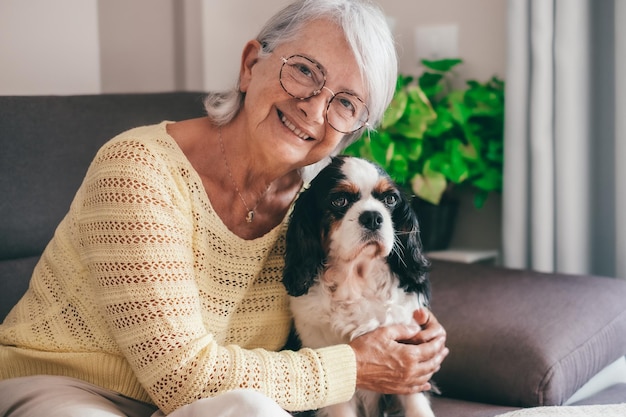 Smiling senior woman sitting on sofa at home while hugging her cavalier king charles spaniel dog