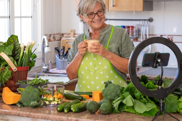 笑顔の年配の女性は、オンラインベジタリアン料理のレッスンに続いてコーヒーブレイクを取ります。野菜だらけの家庭用キッチンテーブル