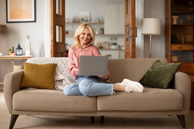 Smiling senior woman using laptop sitting on comfy sofa indoor