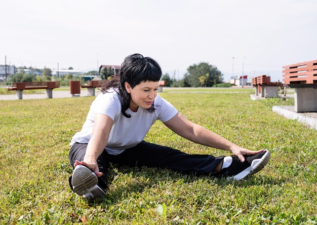 Sorridente donna senior in fase di riscaldamento stretching seduto sull'erba nel parco