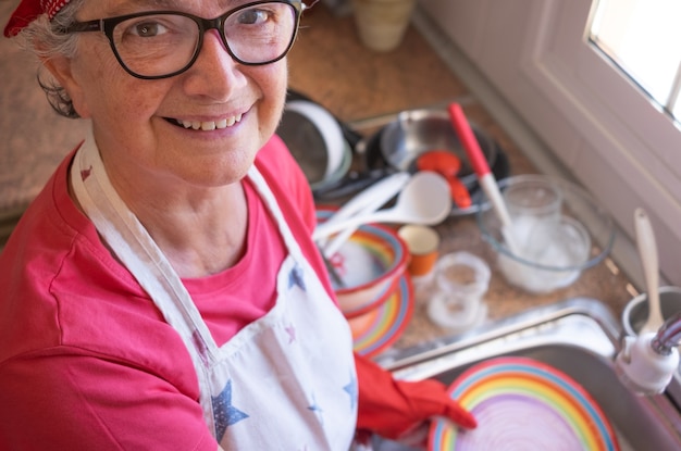 Smiling senior woman washing crockery in front to the window, wearing red gloves and bandana