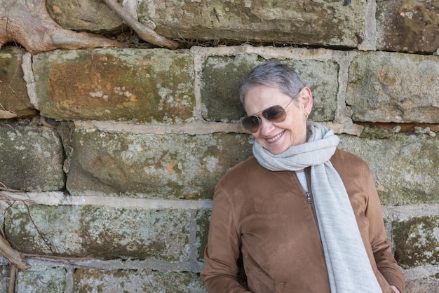 Photo smiling senior woman wearing sunglasses and jacket standing against wall