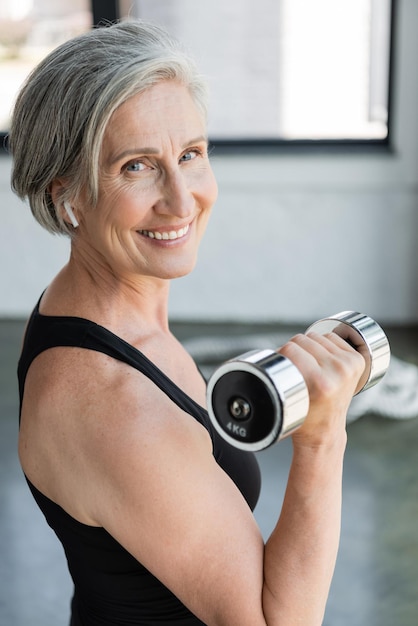 smiling senior woman in wireless earphone listening music and working out with dumbbell in gym