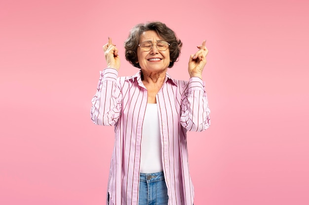 Photo smiling senior woman with crossed fingers standing isolated on pink background with closed eyes