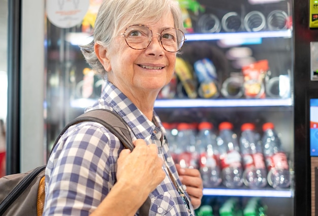 Улыбающаяся пожилая женщина в очках выбирает закуски и напитки на круглосуточном бесплатном автоматическом дистрибьюторе