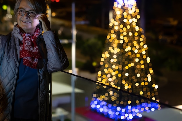 A smiling senior woman with eyeglasses using the cellphone. Outdoor in the night with illuminated Christmas tree behind she. Yellow and blue lights decorations.