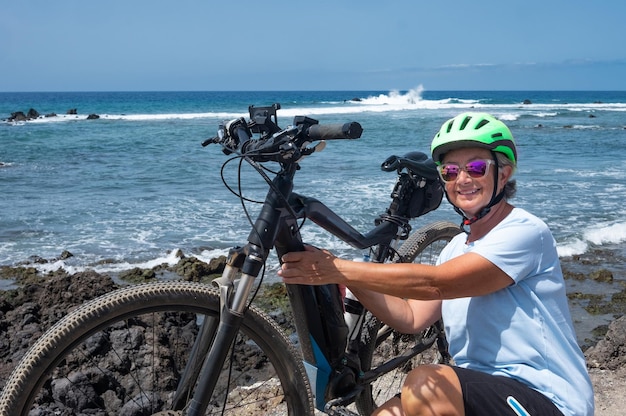 アウトドアと自然と海を楽しむヘルメットと電動自転車を持つ笑顔の年配の女性は、健康的な乗り物の前に彼女の自転車をチェックします