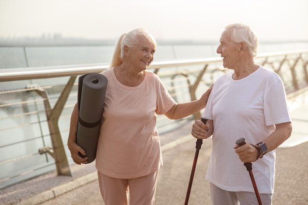 Photo smiling senior woman with mat cheers up man with poles for nordic walking on footbridge