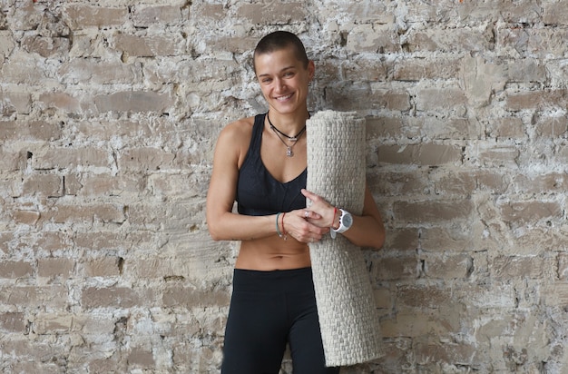 Smiling shorthaired woman in sports wear and yoga mat near brick wall