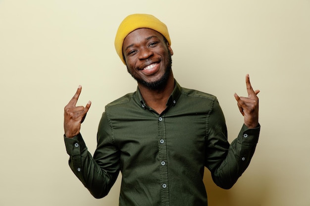 Sorridente che mostra gesto di capra giovane maschio afroamericano con cappello che indossa una camicia verde isolato su sfondo bianco