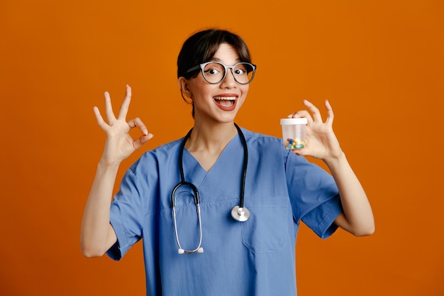 オレンジ色の背景に分離された制服の聴診器を身に着けている錠剤コンテナー若い女性医師を保持している大丈夫ジェスチャーを示す笑顔