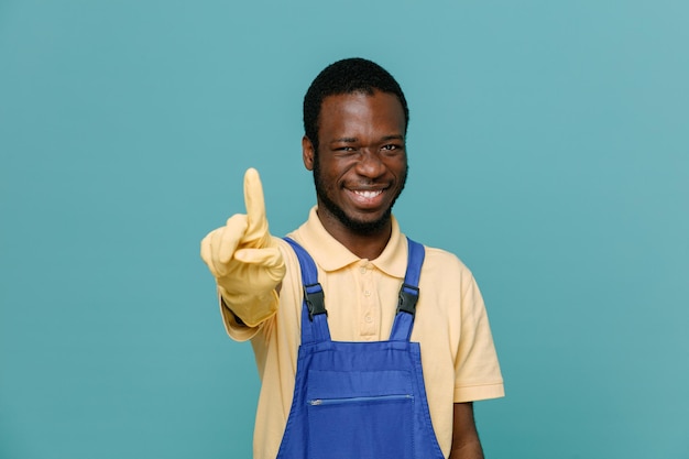 Sorridente che mostra un giovane maschio pulitore afroamericano in uniforme con guanti isolati su sfondo blu