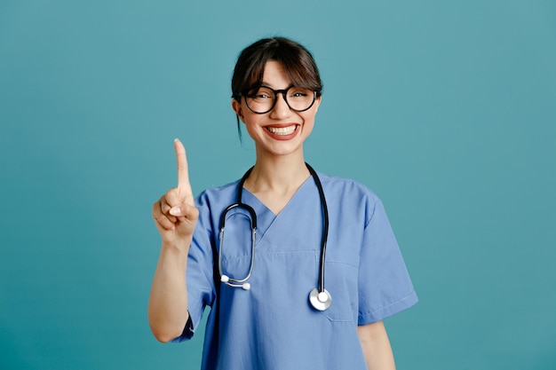 Sorridente che mostra una giovane dottoressa che indossa uno stetoscopio fitth uniforme isolato su sfondo blu