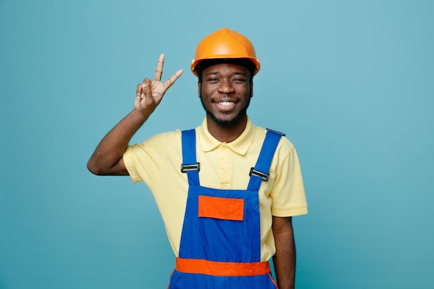 smiling showing peace gesture young african american builder in uniform isolated on blue background