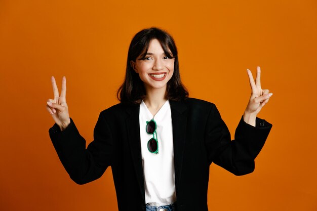 Smiling showing peace gesture young beautiful female wearing black jacket isolated on orange background