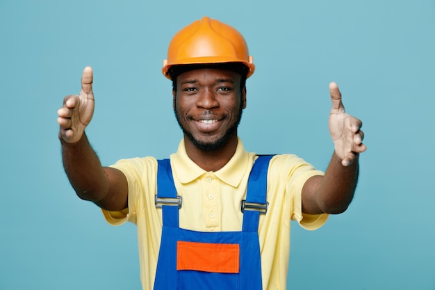 Sorridente che mostra le dimensioni del giovane costruttore afroamericano in uniforme isolato su sfondo blu