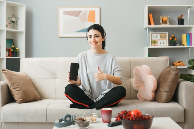 Smiling showing thumb up young girl holding phone sitting on sofa behind coffee table in living room