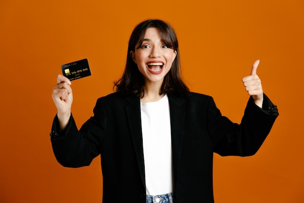 Smiling showing thumbs up holding card young beautiful female wearing black jacket isolated on orange background