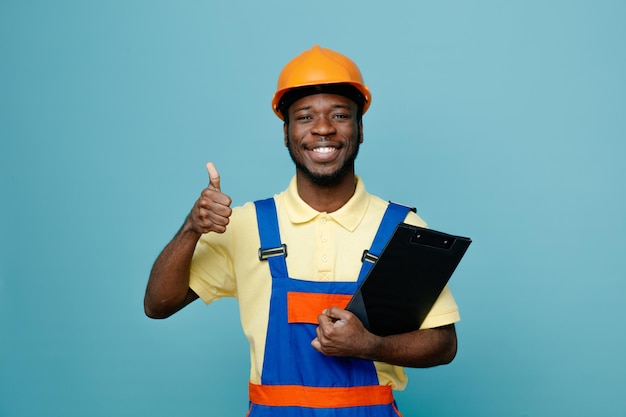 Smiling showing thumbs up young african american builder in uniform holding clipboard isolated on blue background