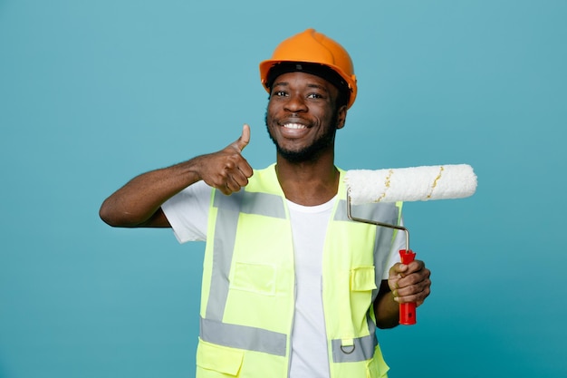 Smiling showing thumbs up young african american builder in uniform holding roller brush isolated on blue background