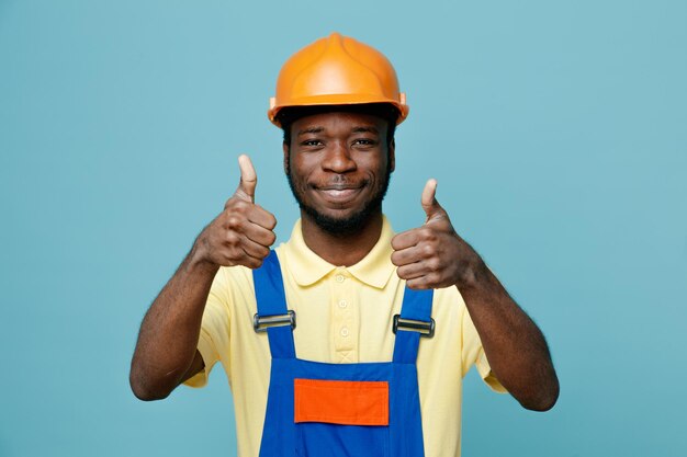 Smiling showing thumbs up young african american builder in uniform isolated on blue background