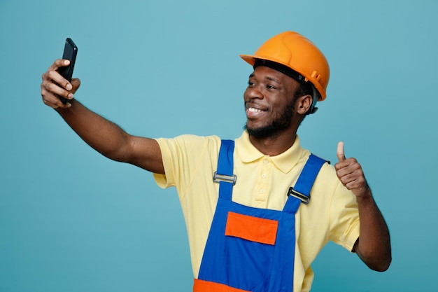 Sorridente mostrando i pollici in su il giovane costruttore afroamericano in uniforme prende un selfie isolato su sfondo blu