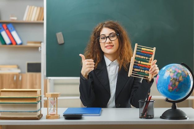 Sorridente che mostra i pollici in su giovane insegnante femminile che indossa occhiali con abaco seduto alla scrivania con strumenti scolastici in classe