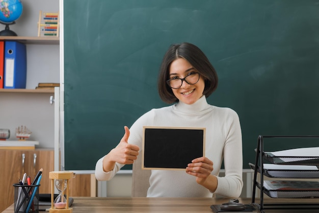 smiling showing thumbs up young female teacher wearing glasses holding mini chalkboard sitting at desk with school tools on in classroom