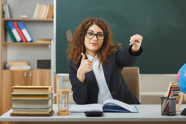 Photo smiling showing thumbs up young female teacher wearing glasses points at camera with pointer sitting at desk with school tools in classroom